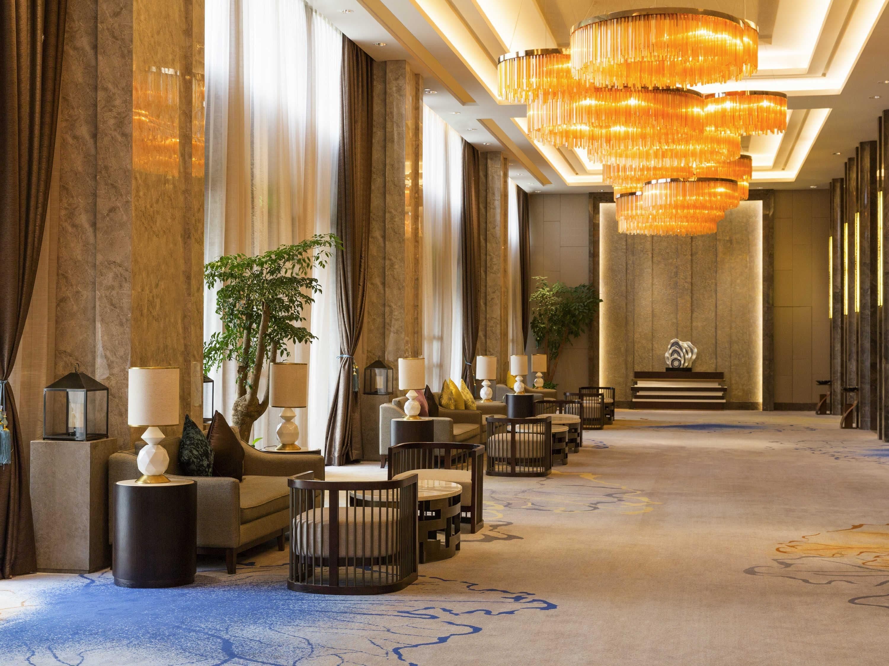 A hotel lobby with a long reception desk, multiple seating areas, and a large chandelier.