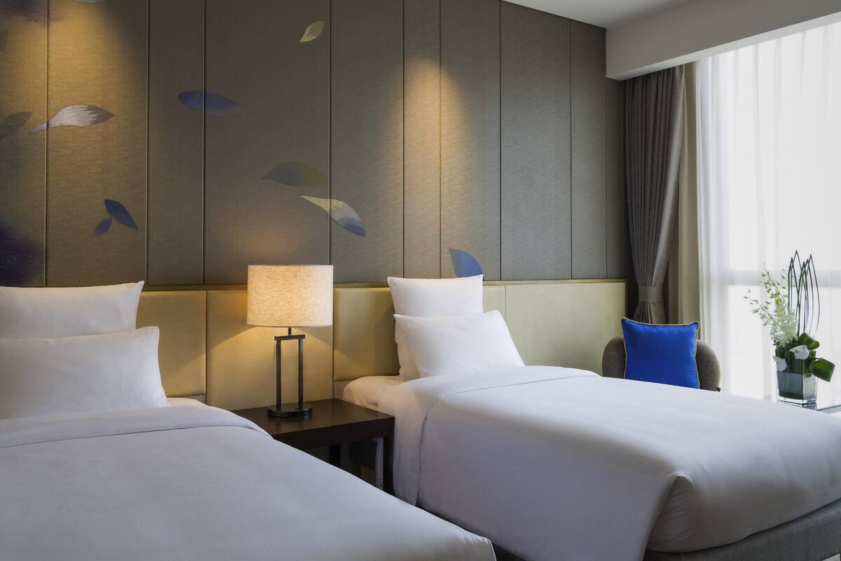 Quarto de hotel com duas camas, mesa de cabeceira com abajur, janela com cortinas e parede com padrão de folhas.