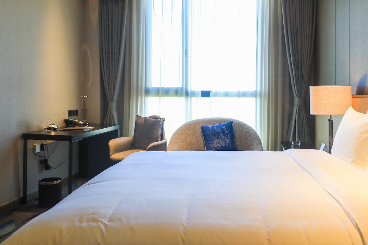 Habitación de hotel con una cama grande, un escritorio con lámpara, una silla y vistas al exterior a través de la ventana.