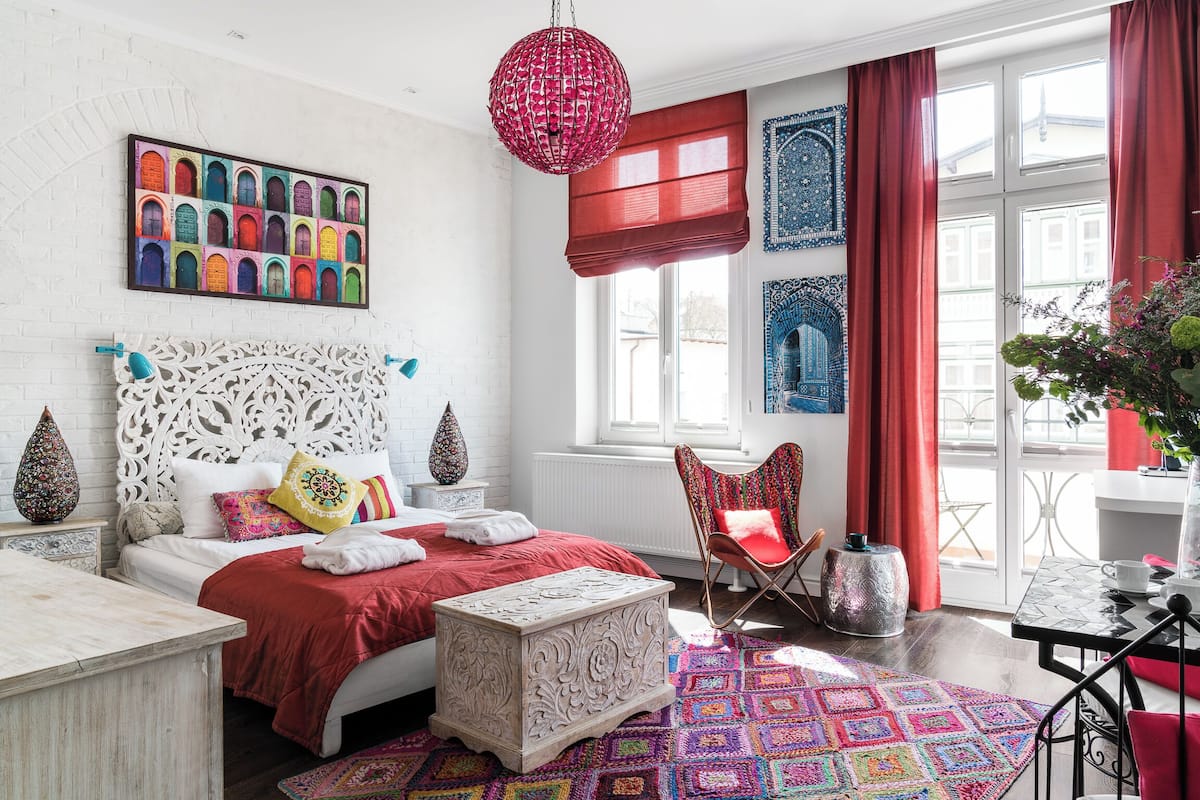 A bedroom with a white brick wall, a colorful door painting, a red pendant light, a patterned rug, and a window with red curtains.