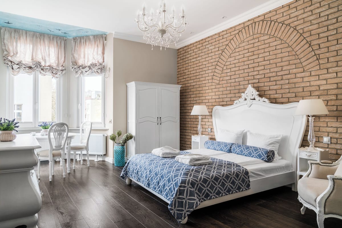 A bedroom with a brick wall, a white bed, a chandelier, and a dining area with chairs and a table.