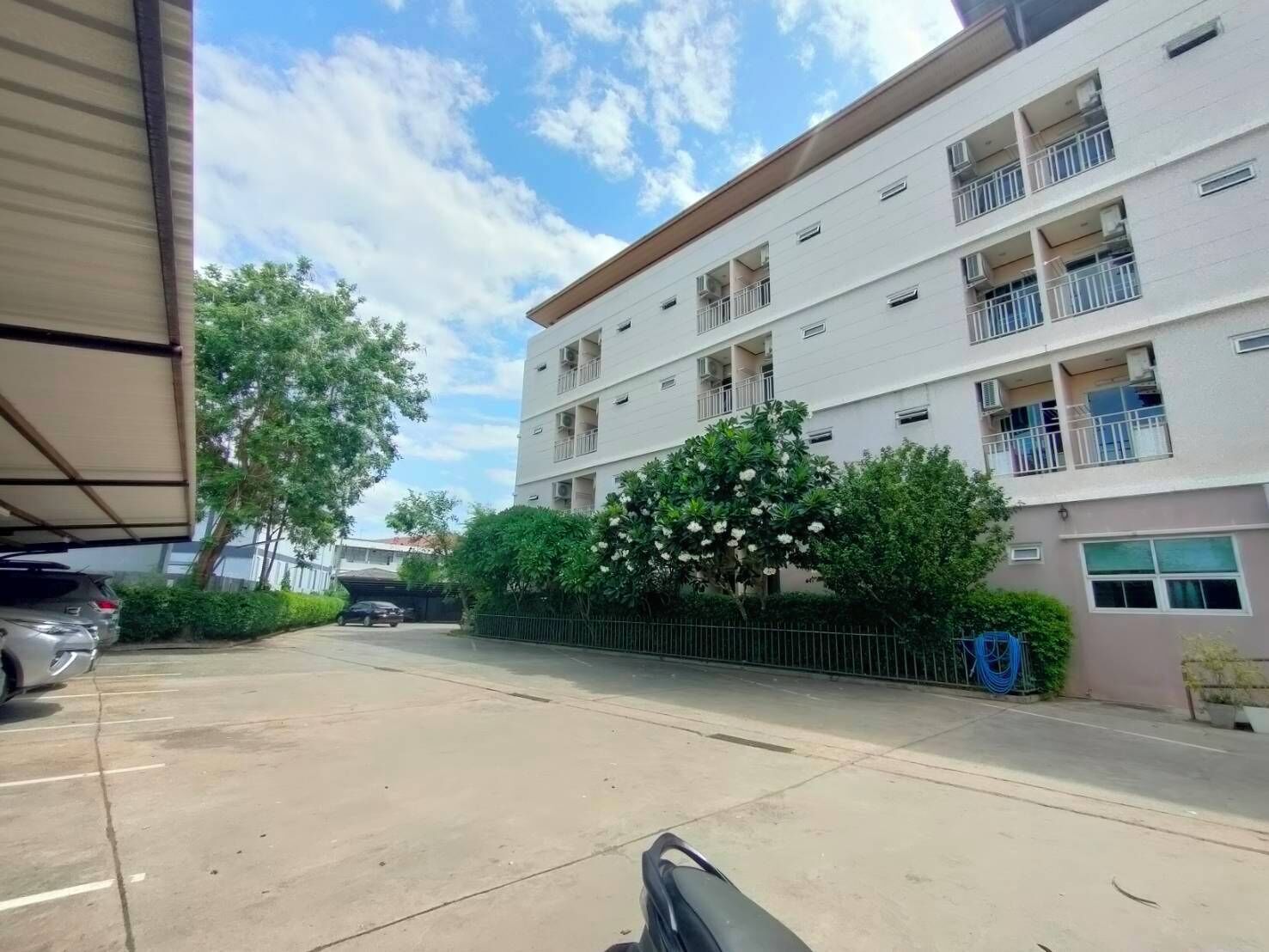 Un edificio de varios pisos con balcones, un estacionamiento y un árbol con flores blancas.