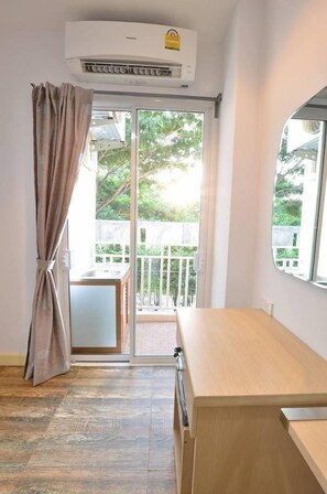 Una habitación con puerta de vidrio corredera que da a un balcón, un escritorio de madera y un equipo de aire acondicionado en la pared.
