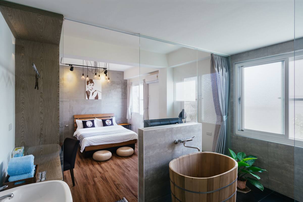 A modern hotel room with a wooden floor, a bed with a headboard, a large window, a wooden bucket, and a bathroom with a bathtub.