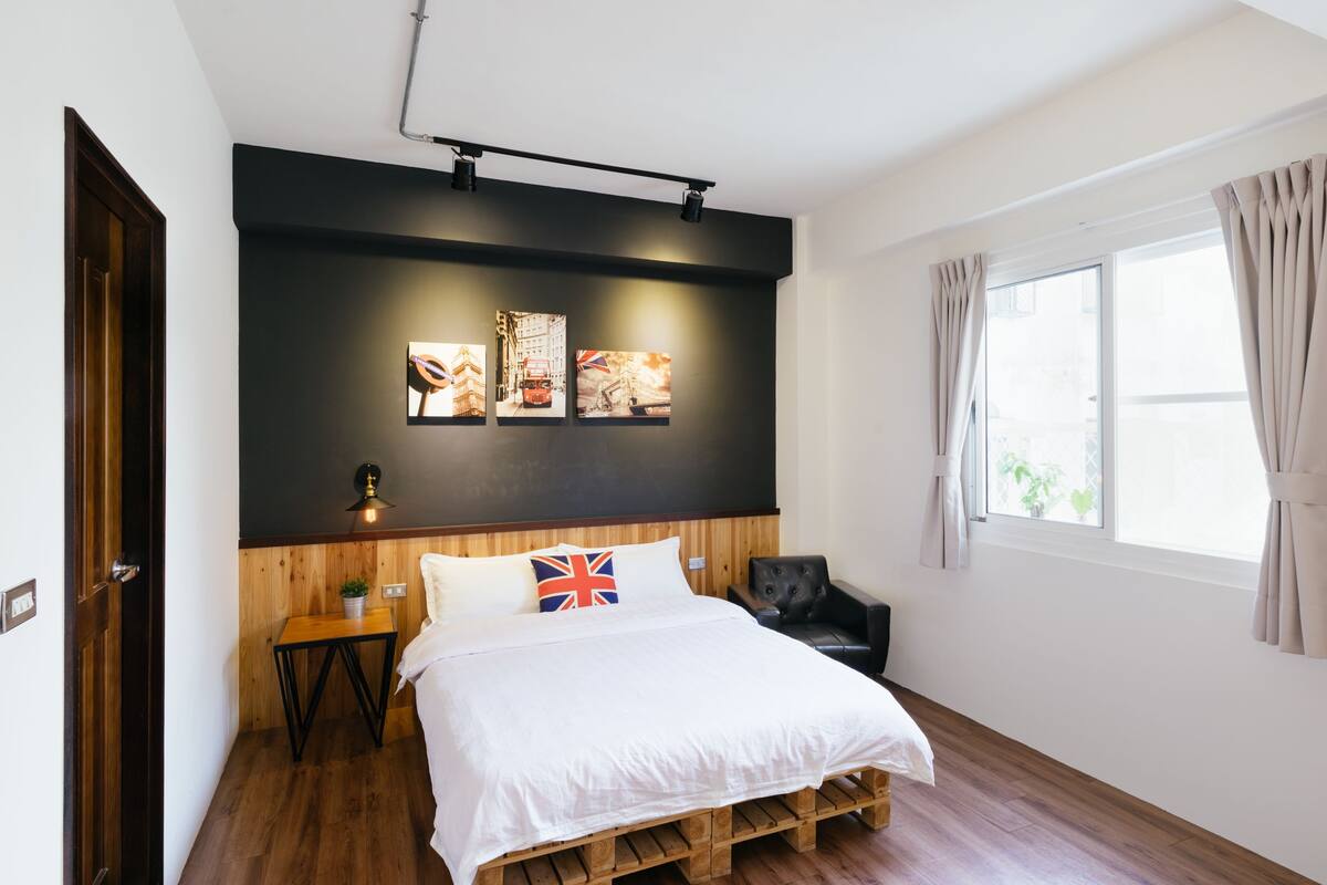 A bedroom with a bed, a small table with a plant, a black chair, and a window with curtains.
