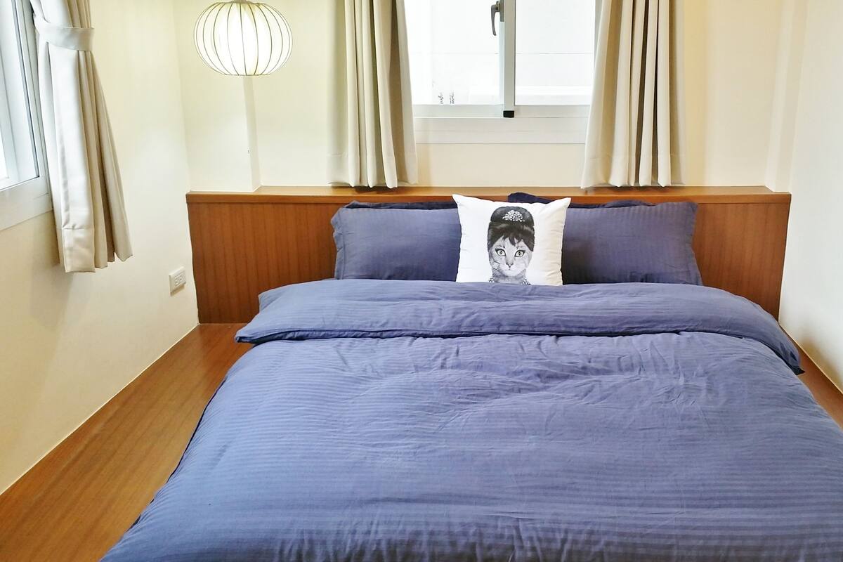 A neatly made bed with a blue comforter and pillows, a wooden headboard, and a window with curtains.