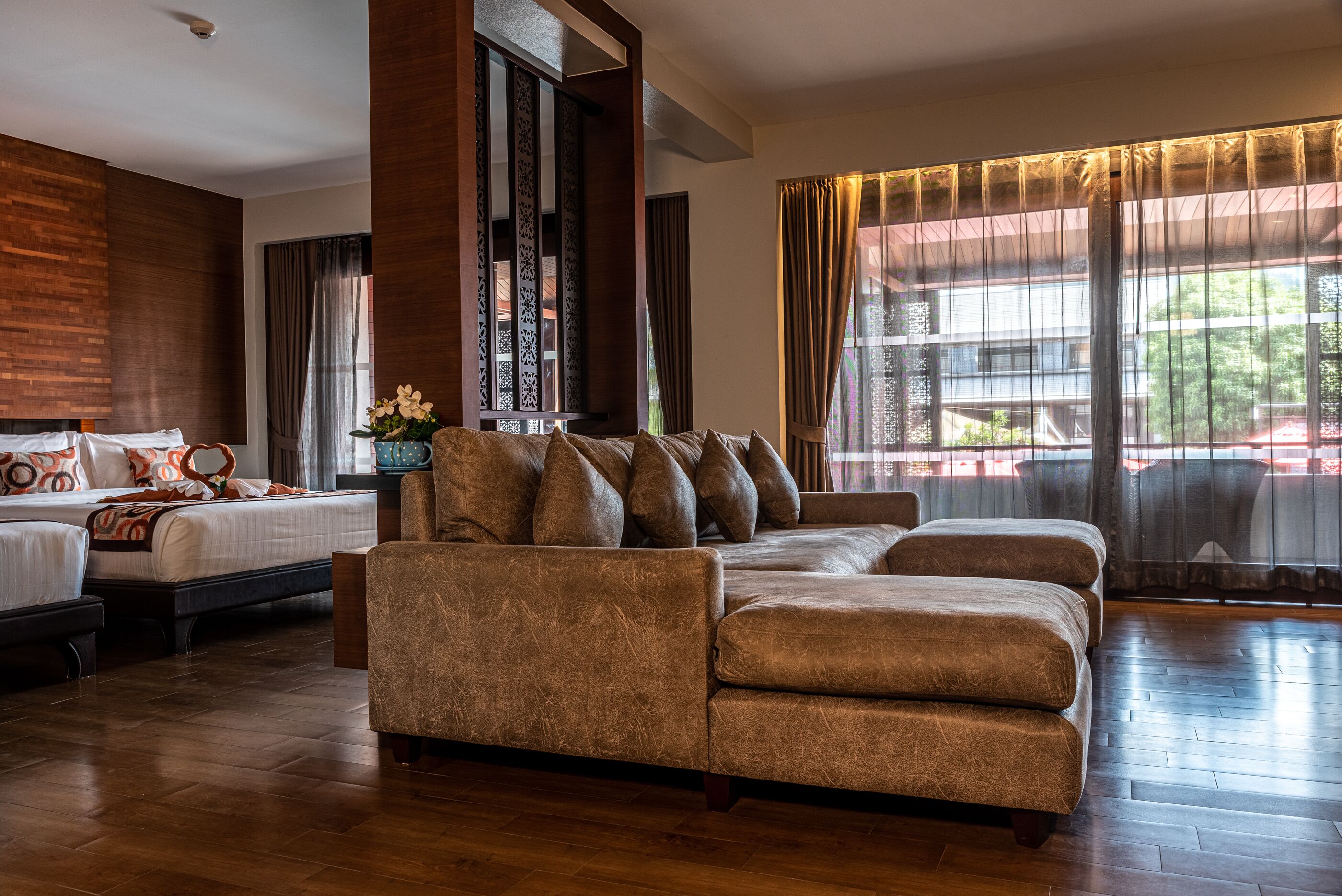 Een hotelkamer met een grote bank, een bed met veel kussens, een klein tafeltje met een bloevas en grote ramen voorzien van gordijnen.