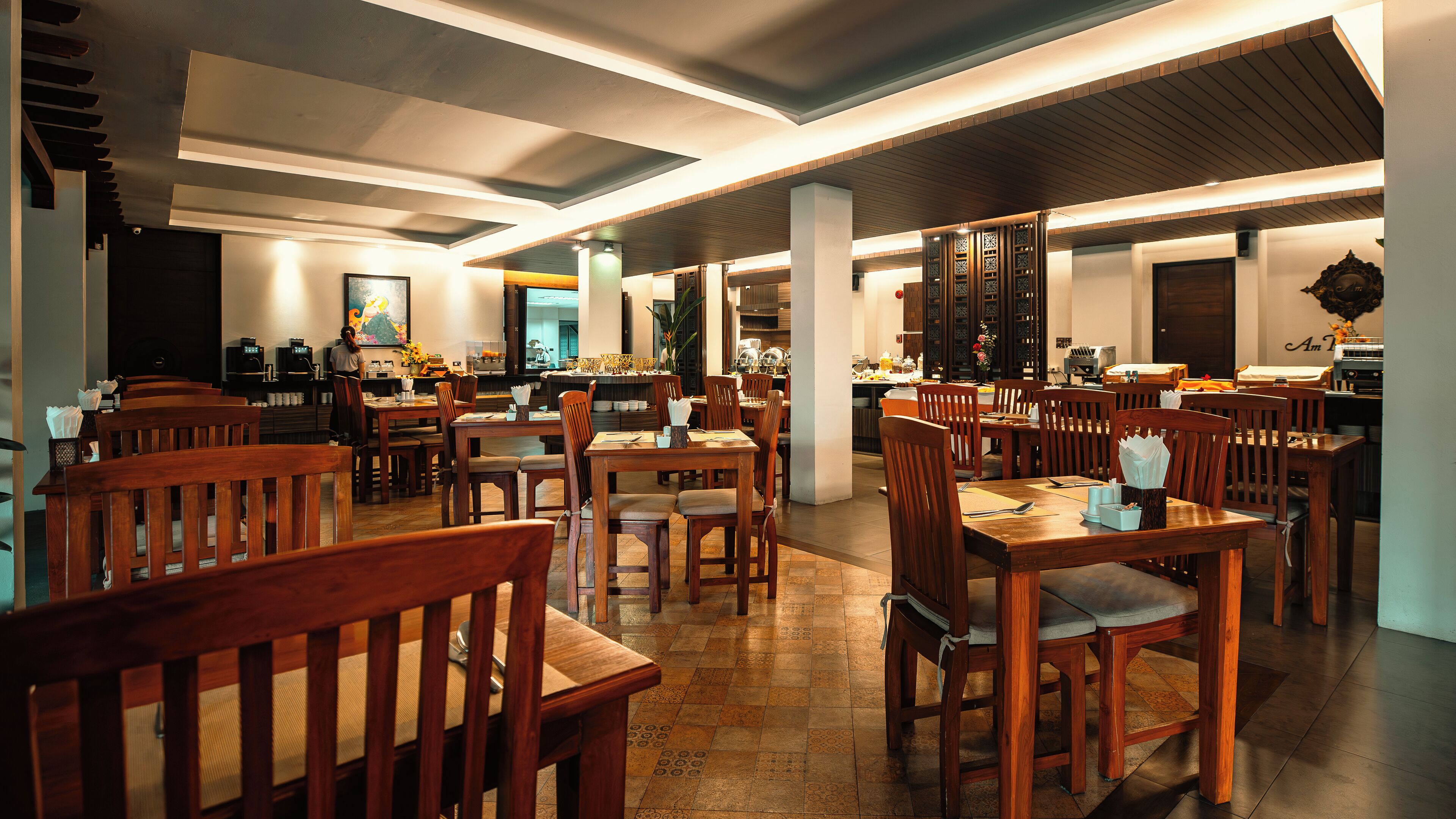 A spacious restaurant with wooden tables and chairs, a kitchen in the background, and a modern ceiling design.