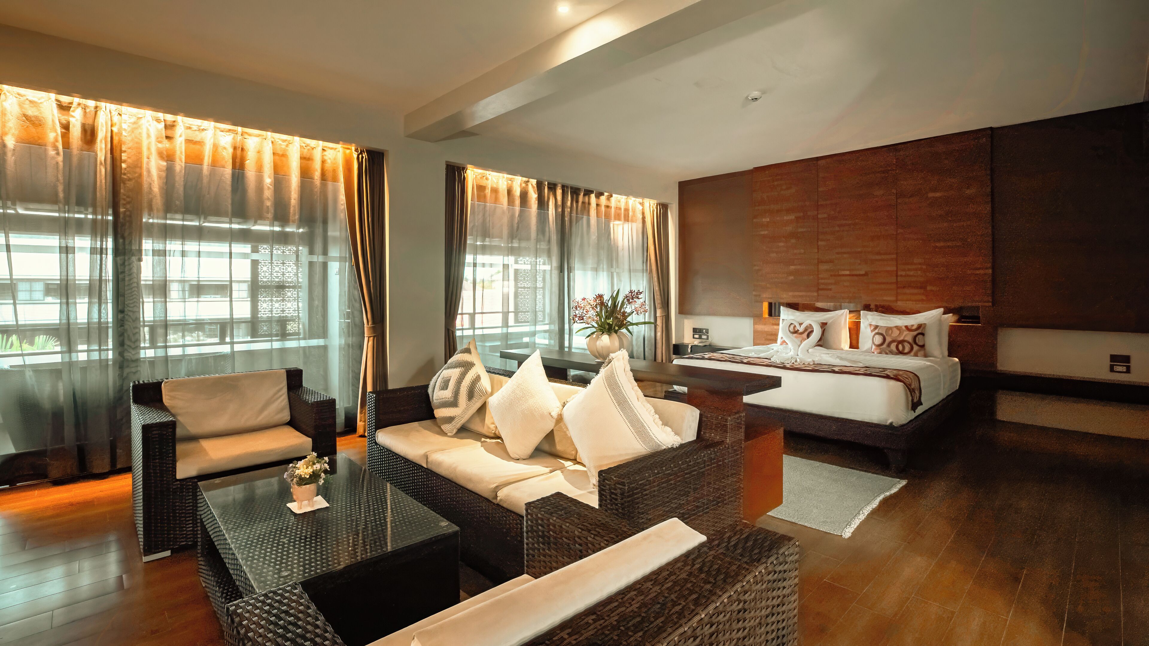 A modern hotel room with a large bed, a sofa, and a coffee table. There are curtains on the windows, and a plant on the coffee table.