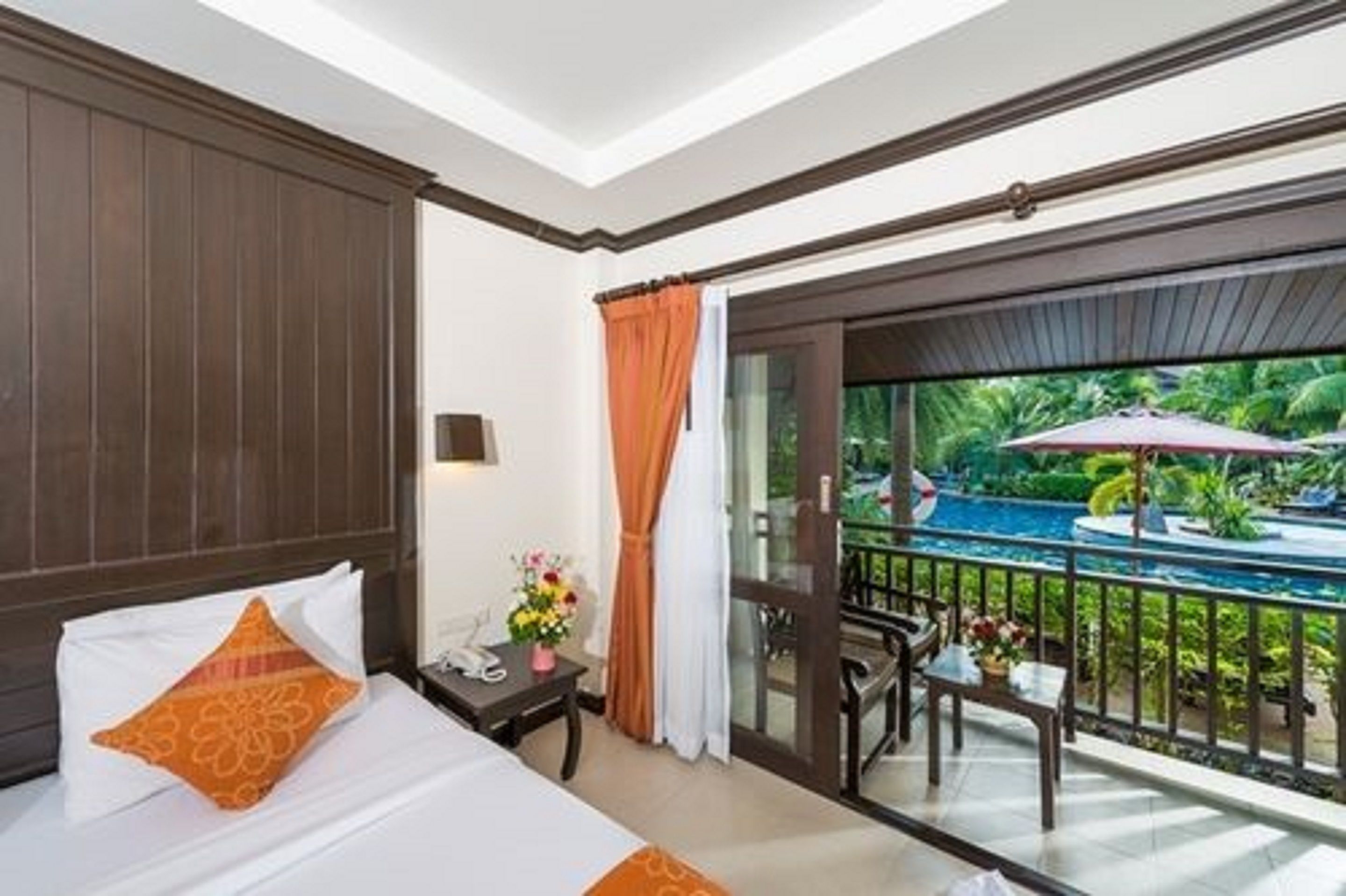 A hotel room with a bed, a nightstand, a table with a vase of flowers, and a view of a swimming pool and tropical garden.