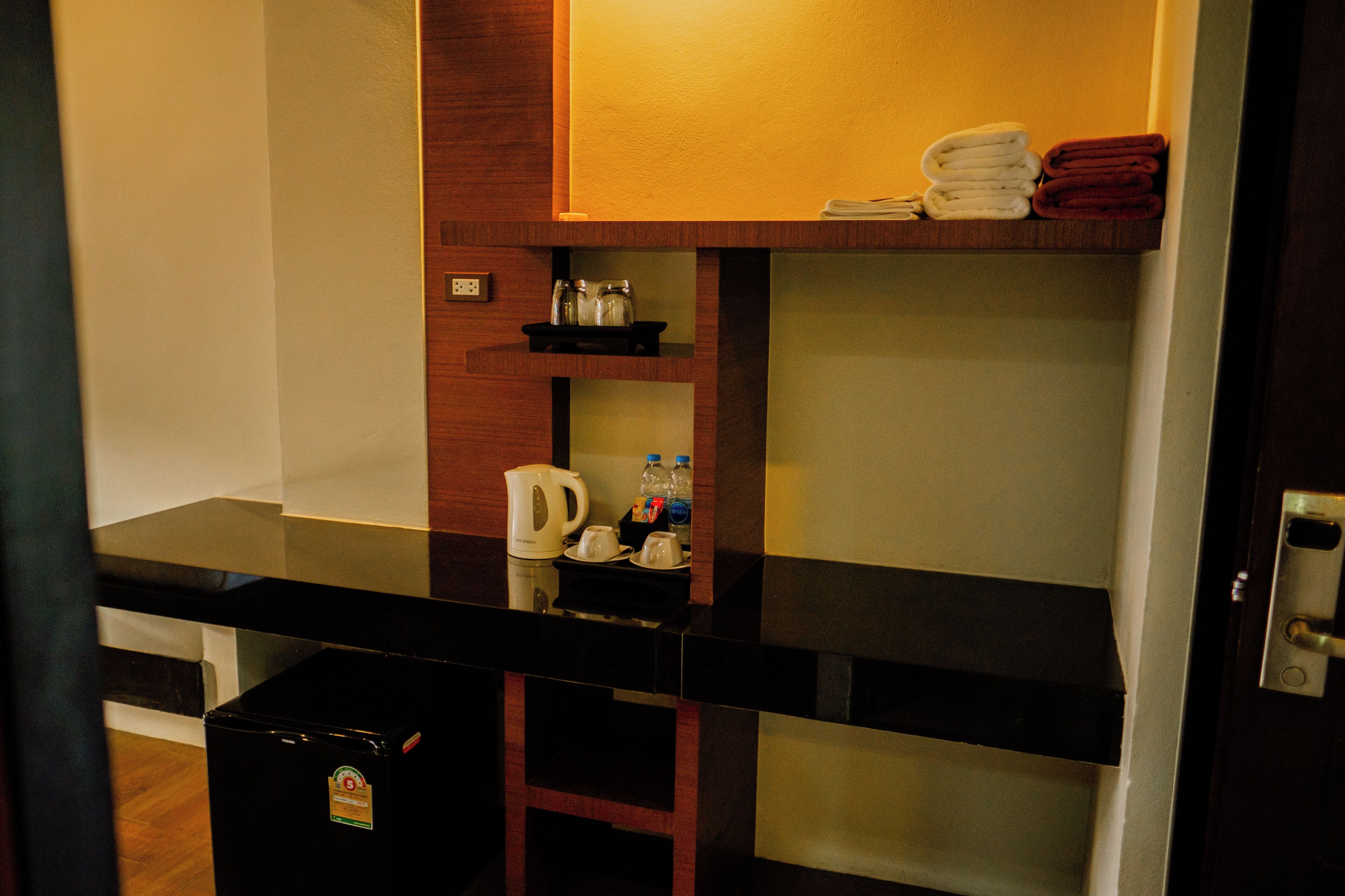 A hotel room with a kitchenette, a coffee maker, a kettle, and a small shelf with towels and bottles.