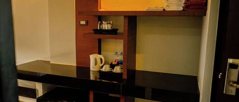 Habitación de hotel con kitchenette, cafetera, hervidor de agua y un pequeño estante con toallas y botellas.