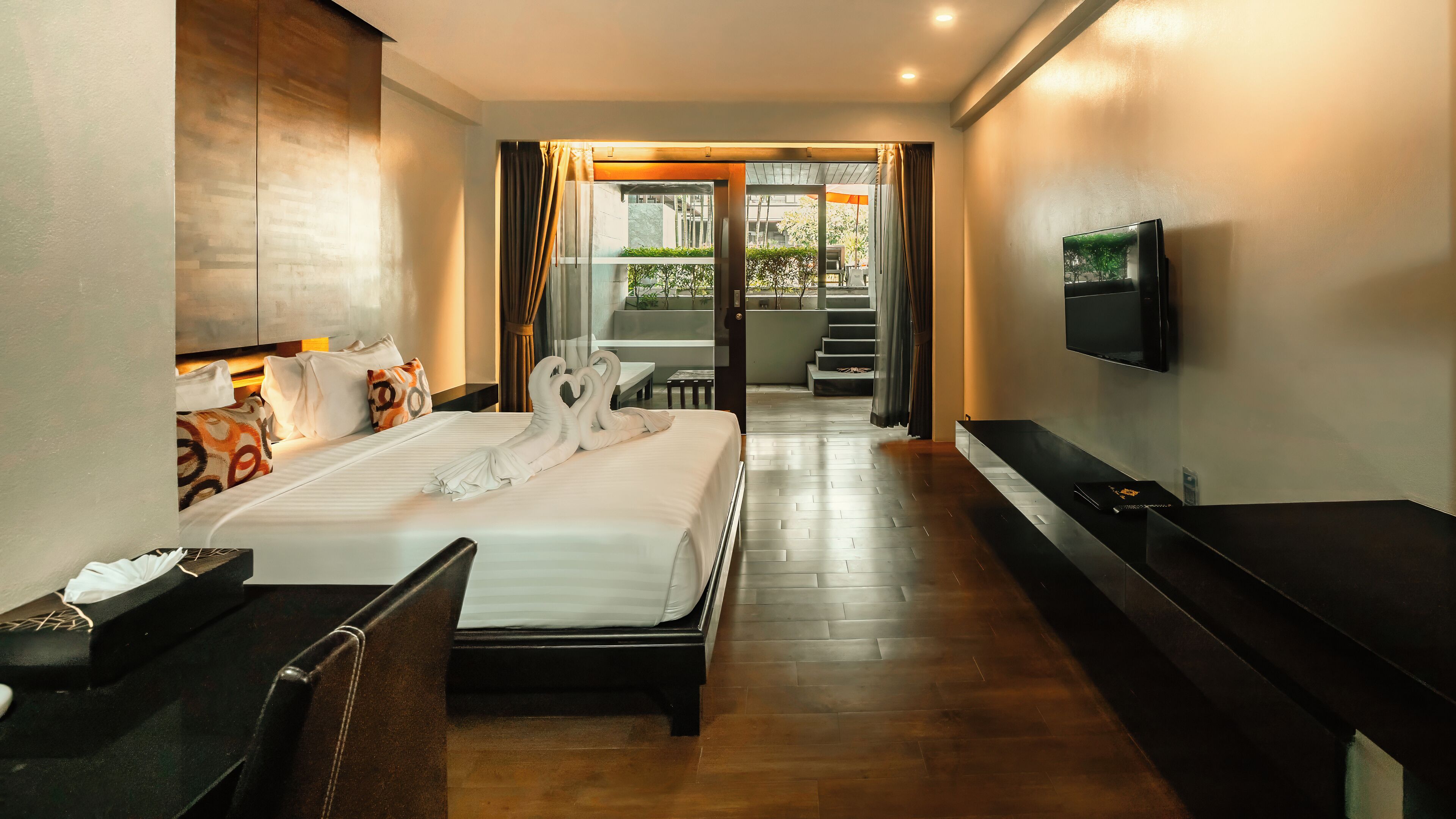 A hotel room with a large bed, a TV mounted on the wall, a desk, and a chair. There is a staircase leading to an outdoor area.