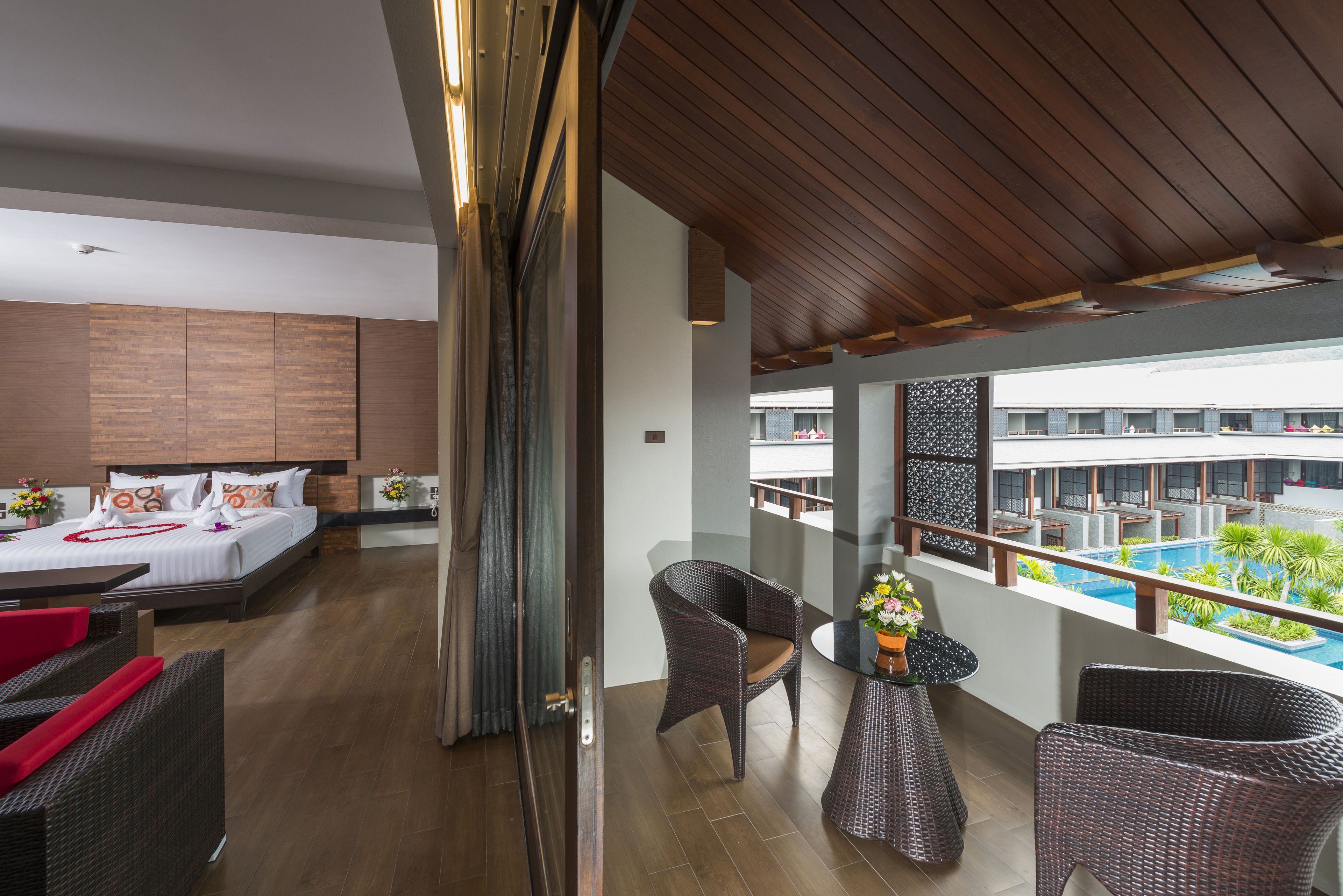 A modern hotel room with a balcony, a bed with white linens, a dining area with wicker furniture, and a view of a pool and buildings.