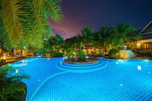 A large, illuminated swimming pool surrounded by palm trees and a building with a curved roof. - Am Samui Palace (Koh Samui)