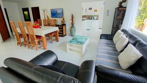A modern living room with a black leather sofa, wooden dining chairs, a glass coffee table, and a TV mounted on the wall.