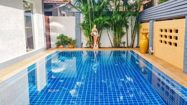 A swimming pool with blue tiles, surrounded by potted plants and a yellow wall with ventilation holes.