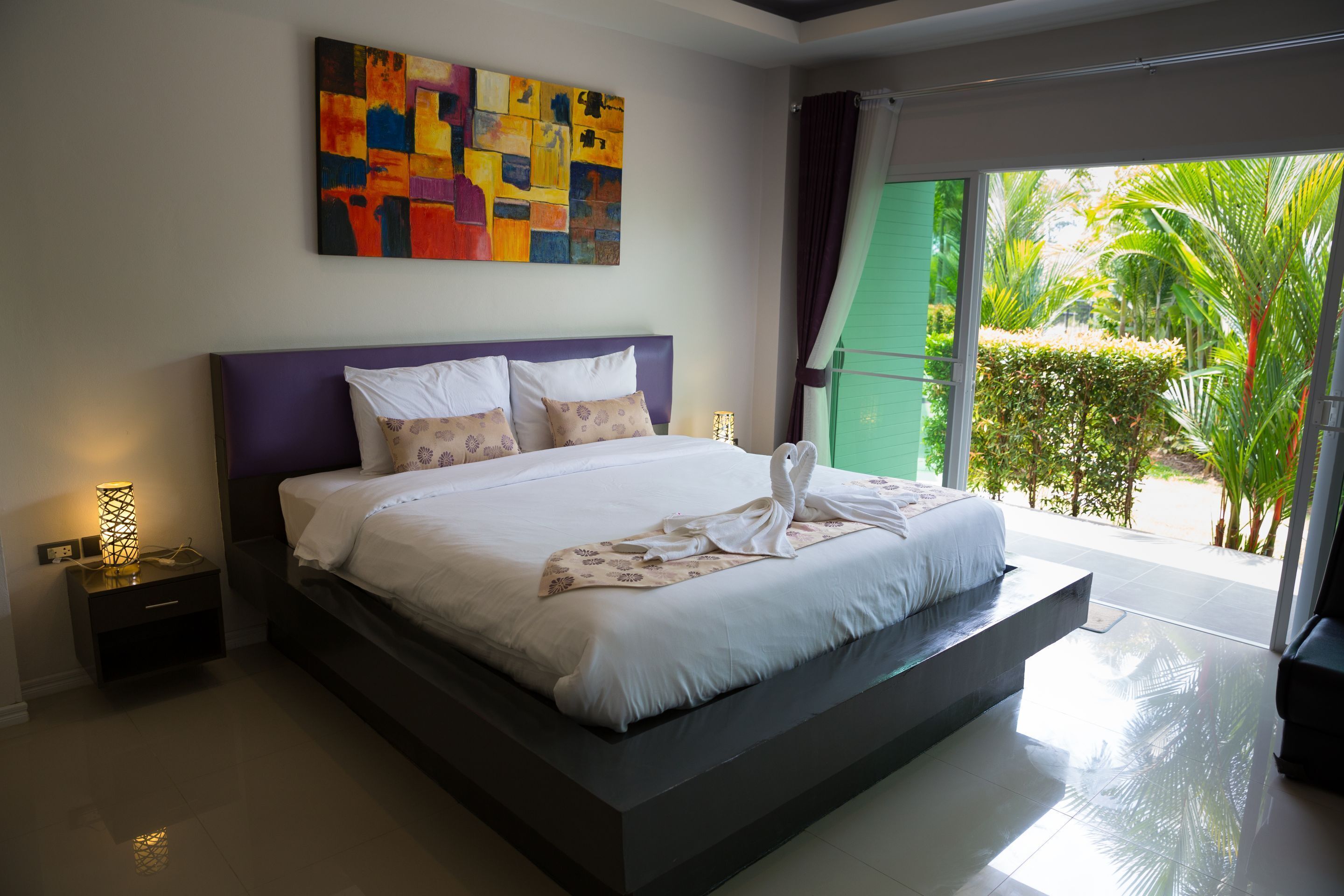 A modern bedroom with a large bed, a colorful abstract painting, and a view of greenery outside.