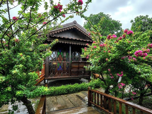 Une construction traditionnelle en bois, dotée d’un balcon, et entourée d’une végétation luxuriante et de fleurs roses.