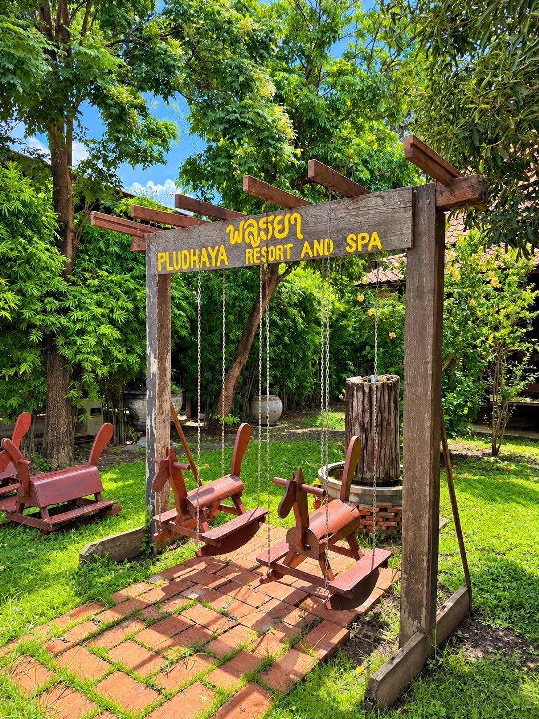 A wooden sign with 'PLUDHAYA RESORT AND SPA' written on it, surrounded by greenery and a swing set.