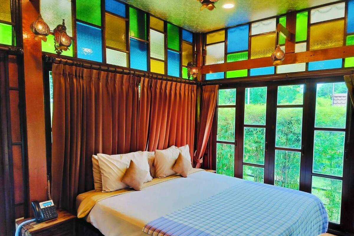 Una habitación con cama, cortinas, mesita de noche con teléfono y ventanas amplias con vista a zonas verdes.