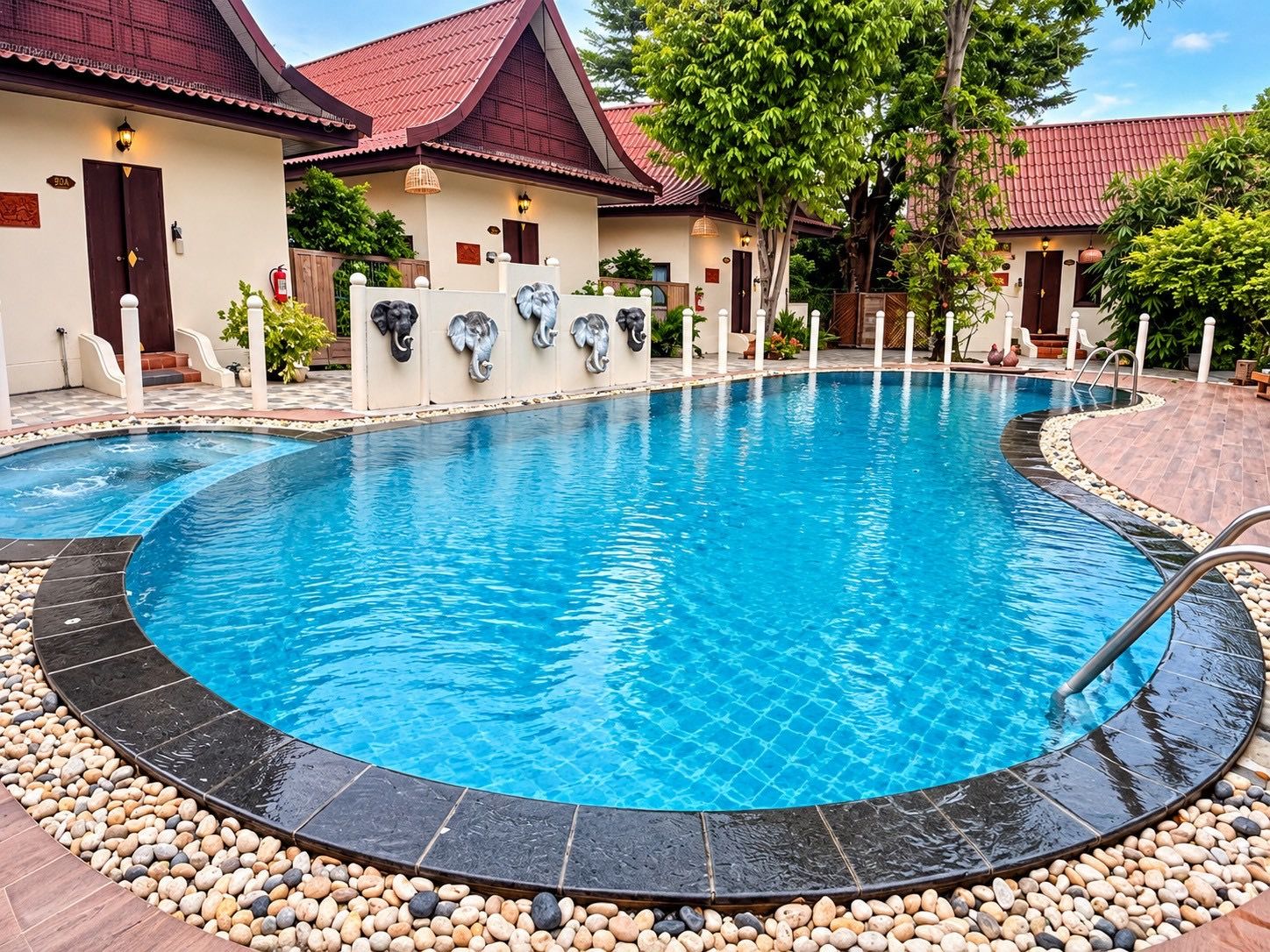 A swimming pool with a curved edge, surrounded by a pebble border, and adjacent to a building with a red-tiled roof.