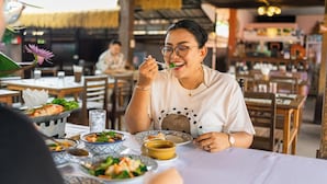Un restaurante con mesas y sillas, una persona comiendo y un techo de paja.
