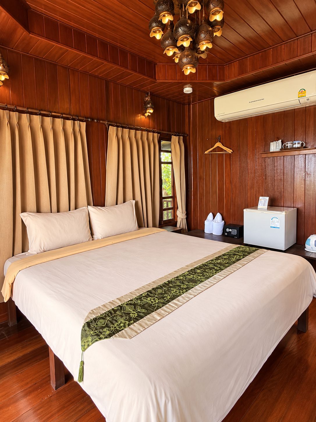 A hotel room with a large bed, wooden panel walls, a ceiling light fixture, an air conditioning unit, and a bedside table with a radio and a cup.