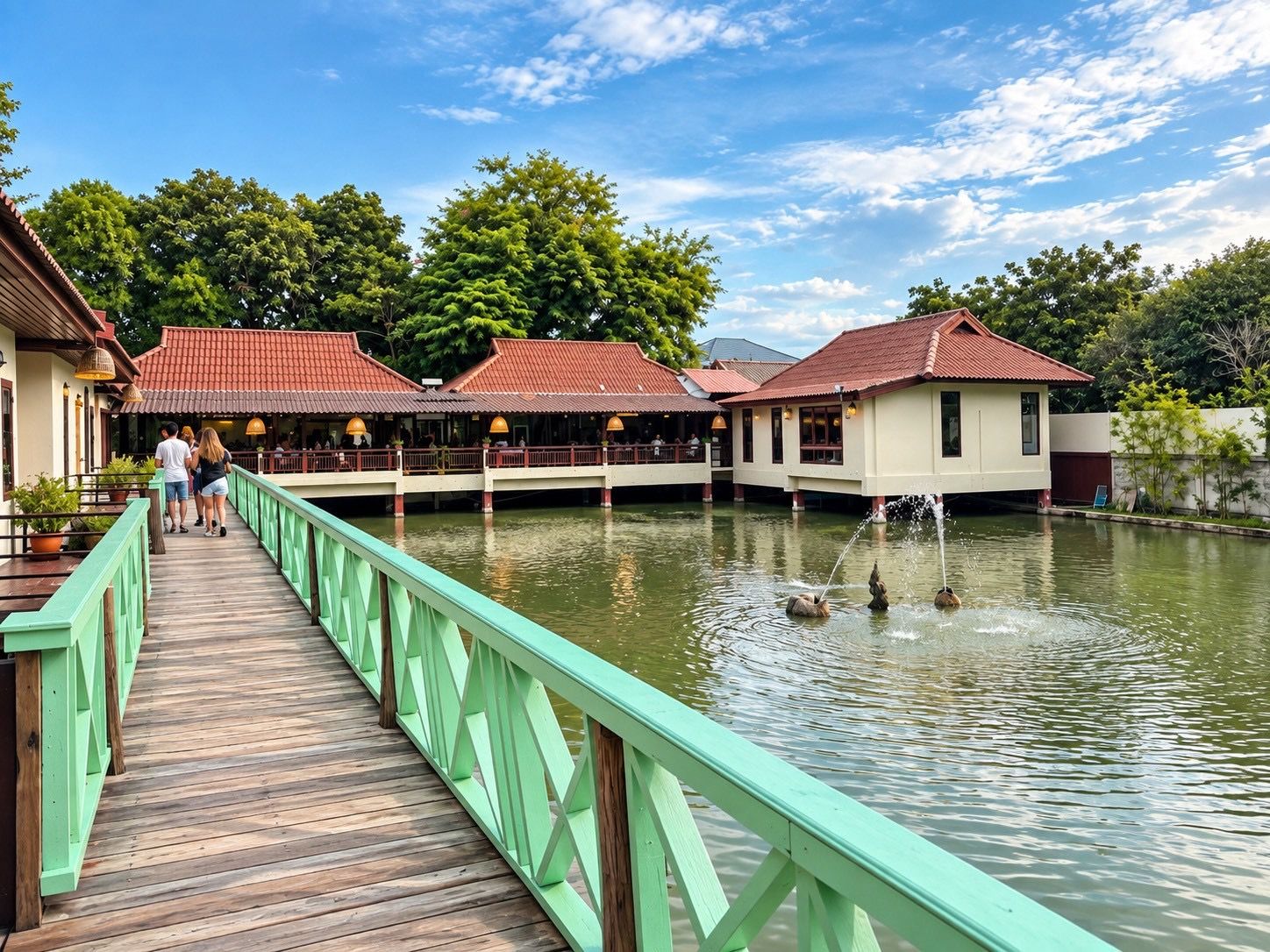 A wooden boardwalk leading to a building with a red roof, over a body of water with a fountain.