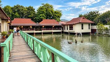 A wooden boardwalk leading to a building with a red roof, over a body of water with a fountain.