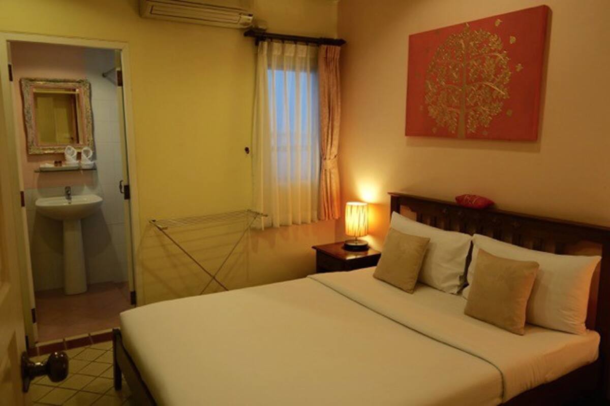 Una camera d'albergo con un letto, un comodino con lampada, una finestra con tende, un bagno con lavandino e specchio, e un'opera d'arte rossa appesa al muro.