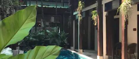 Una piscina con piso de baldosas, rodeada de vegetación y un edificio de varios pisos con balcones.
