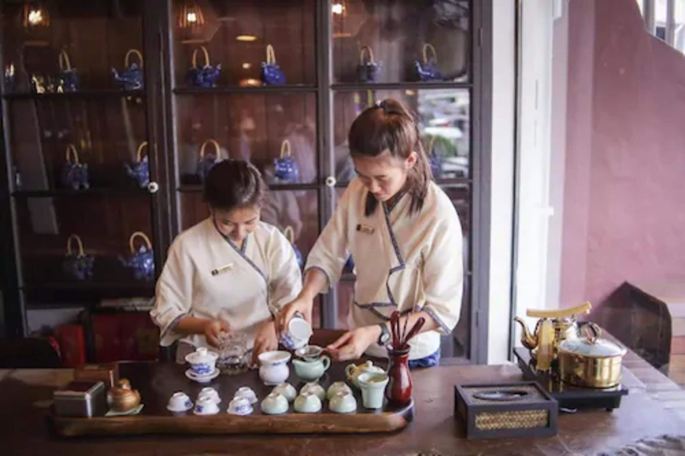 Two individuals serving tea at a table with multiple teapots and cups.