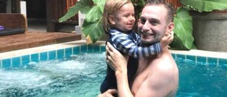 A man and a child in a swimming pool with a large plant and a no smoking sign in the background.