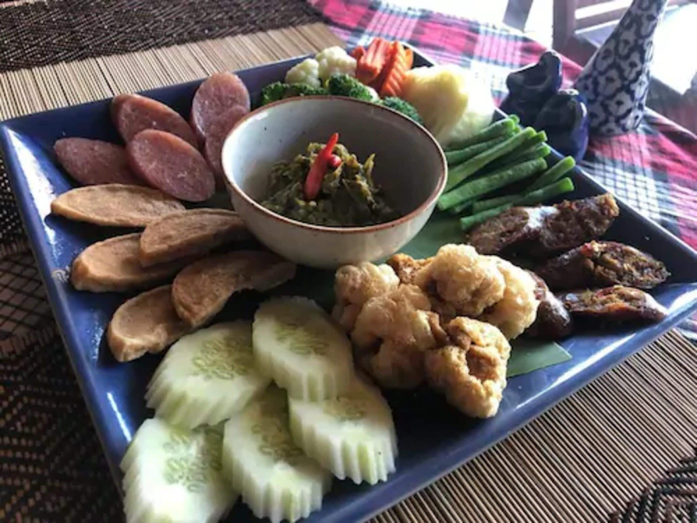 Een vierkant bord met diverse soorten eten, waaronder plakjes worst, gekookte eieren, groene bonen en een schaal salade.