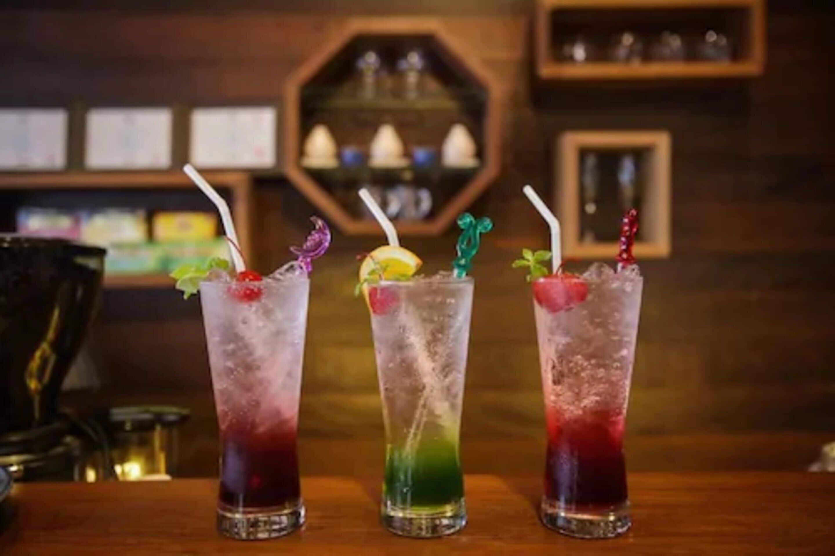Drei hohe Gläser mit Eisgetränken und Trinkhalmen, jedes mit einer anderen farbigen Flüssigkeit und einer Dekoration, auf einem Holztisch in einer Bar.