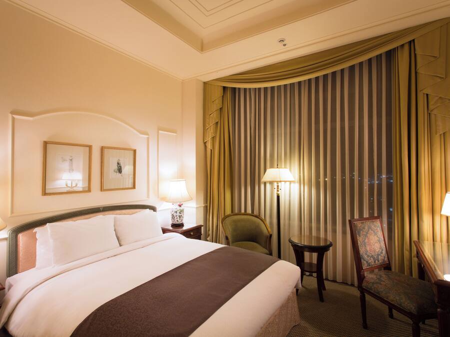 A hotel room with a large bed, two bedside lamps, a chair, a small table, and a window with curtains.