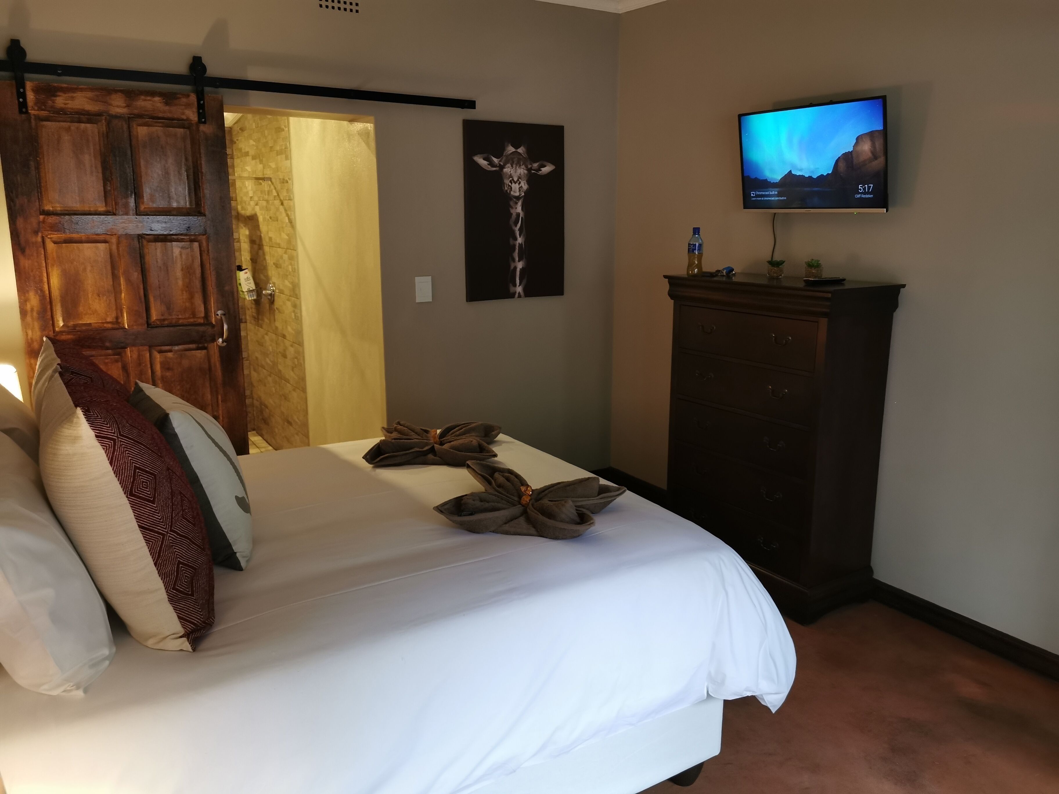 Quarto de hotel com cama, televisão na parede, porta de madeira e quadro com girafa emoldurada.
