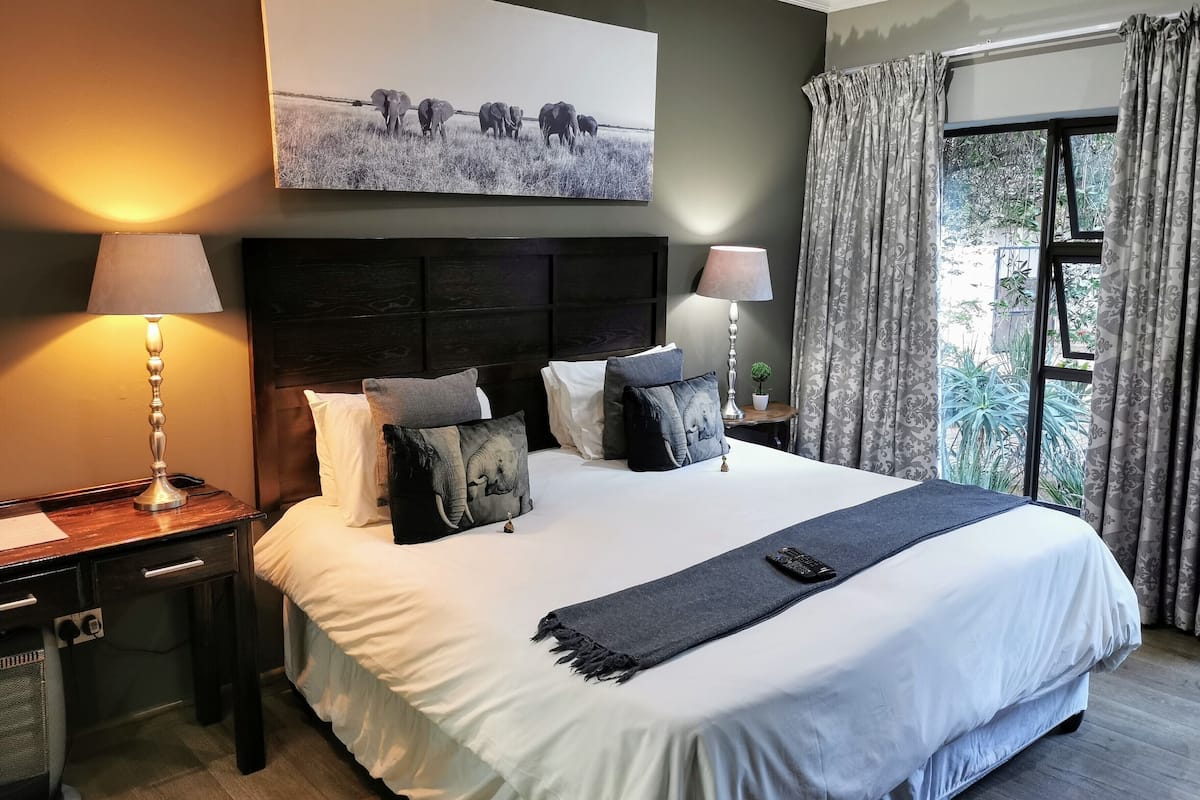 Una cama bien arreglada, con una paleta de colores blanco y gris, flanqueada por dos mesitas de noche con lámparas, un cabecero con un panel decorativo y un cuadro grande sobre la cama.