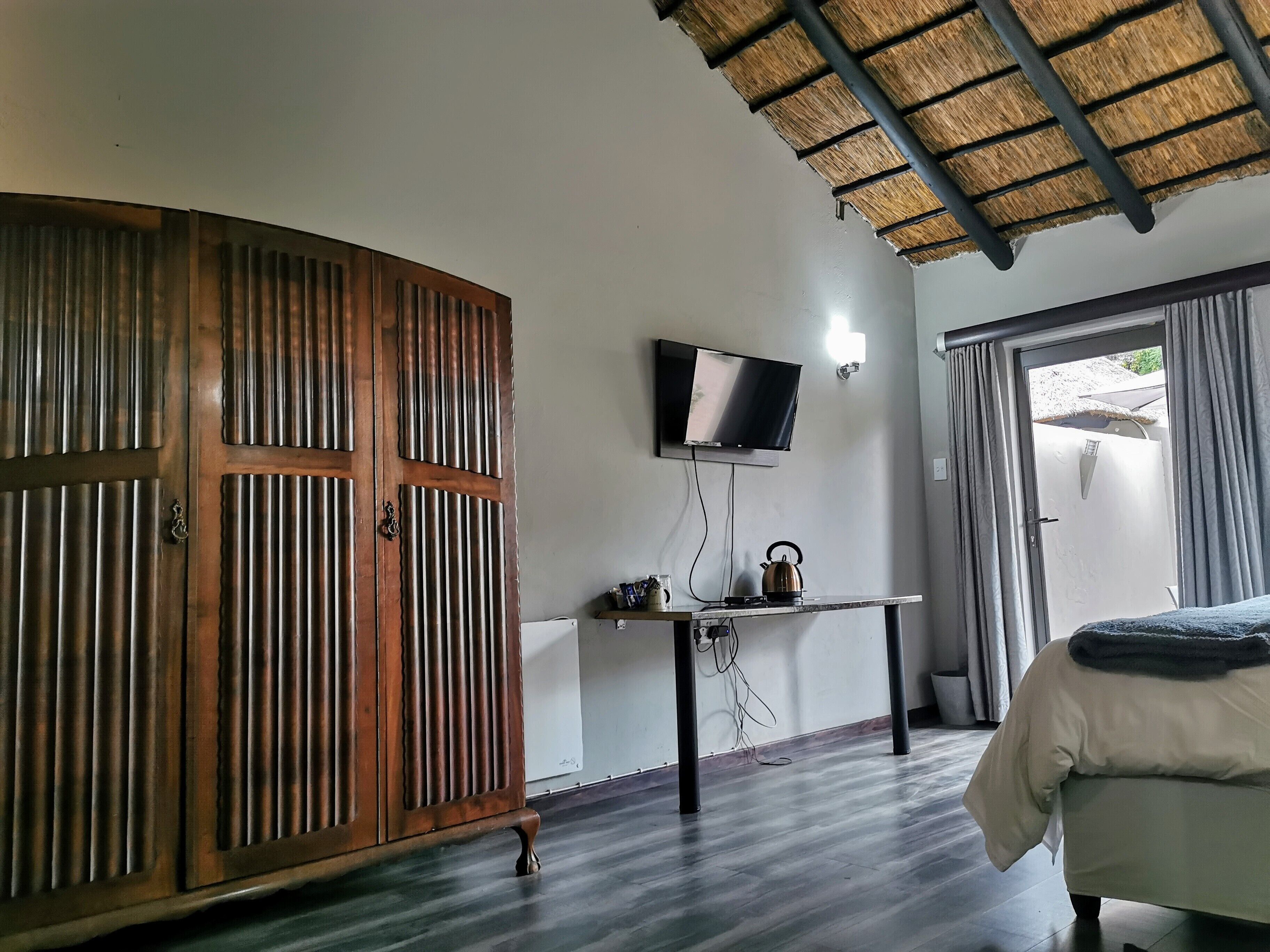 Ein Zimmer mit Holzschrank, Flachbildfernseher an der Wand, Tisch mit Wasserkocher und Bett mit Decke.