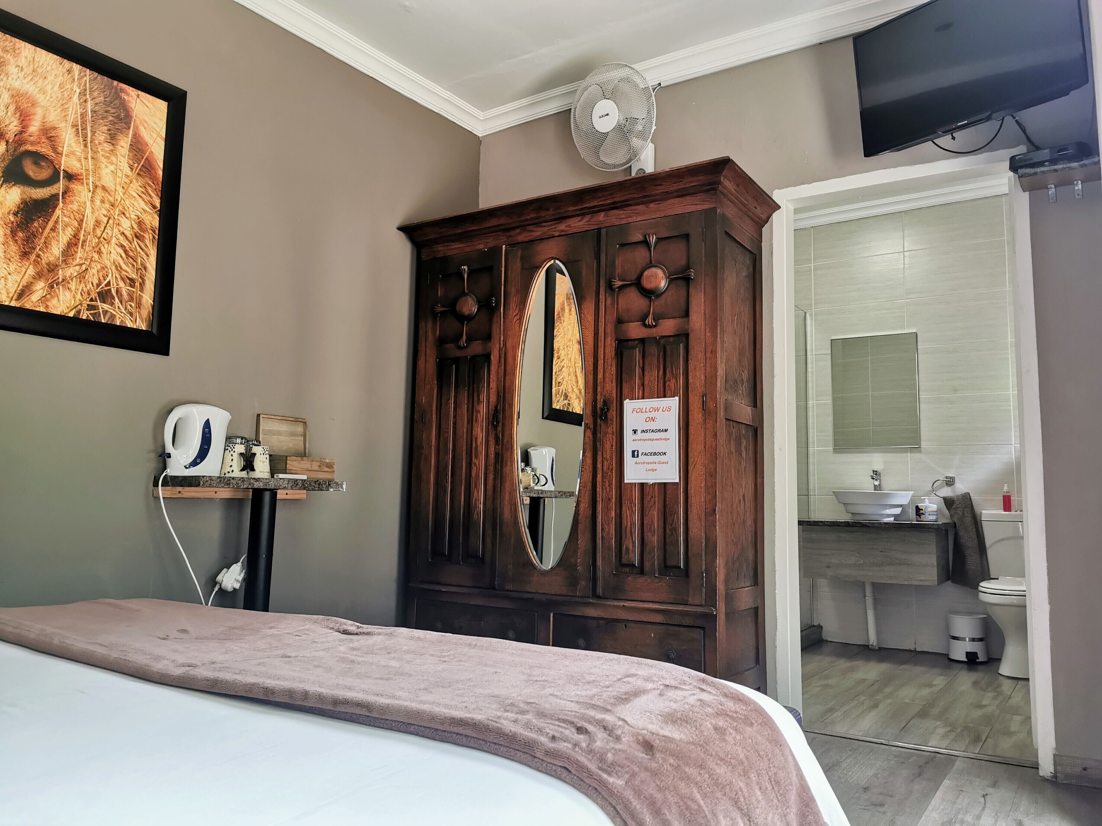 A hotel room with a bed, a wooden wardrobe, a mirror, a fan, a television, and a bathroom visible through an open door.