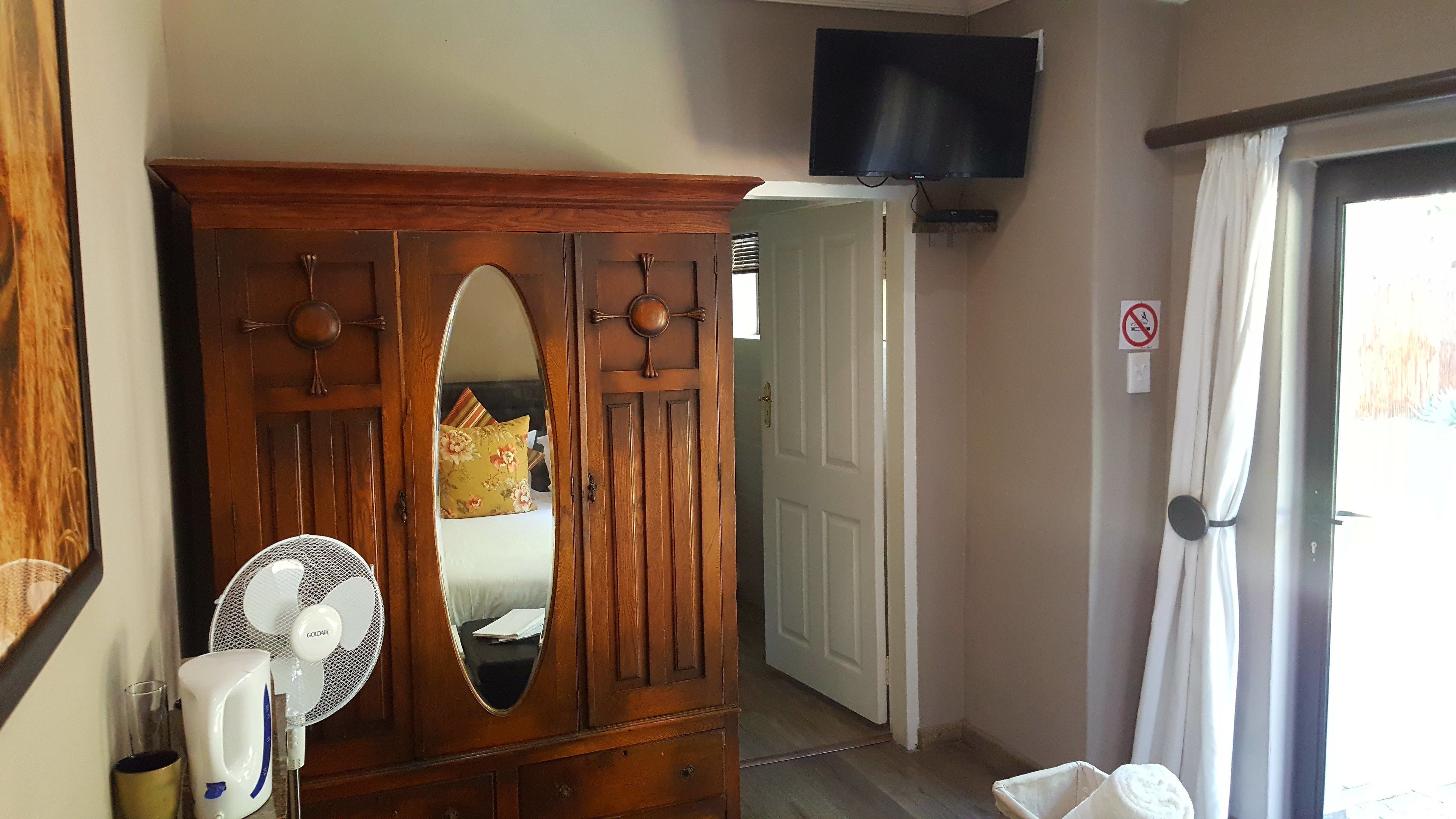 Une chambre avec une armoire en bois, un miroir, un ventilateur, un téléviseur fixé au mur et une porte sur laquelle est apposé un panneau « interdiction de fumer ».