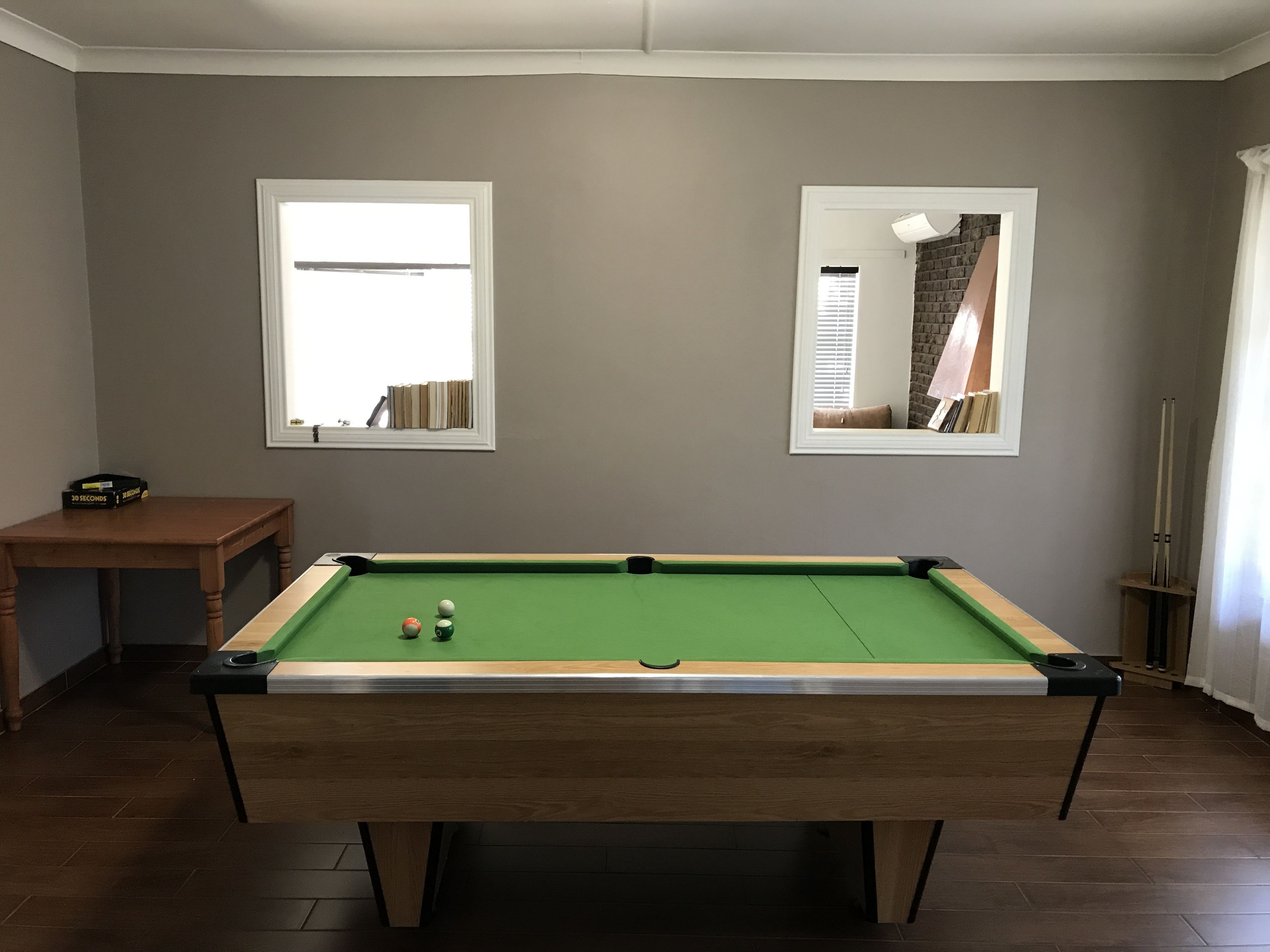 A pool table in a room with two windows, a small table with a box, and a lamp.