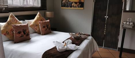 A hotel room with a bed, pillows, a towel, a painting, a wardrobe, and a small shelf with toiletries.