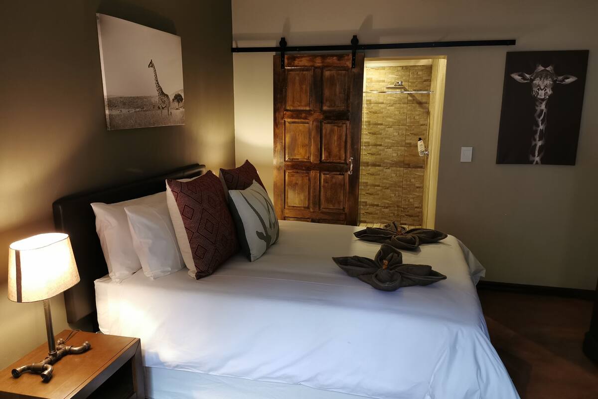 Una cama bien hecha con almohadas blancas y estampadas, una lámpara de noche y una mesita de madera. Se ve una puerta de madera con una pared de piedra al fondo.