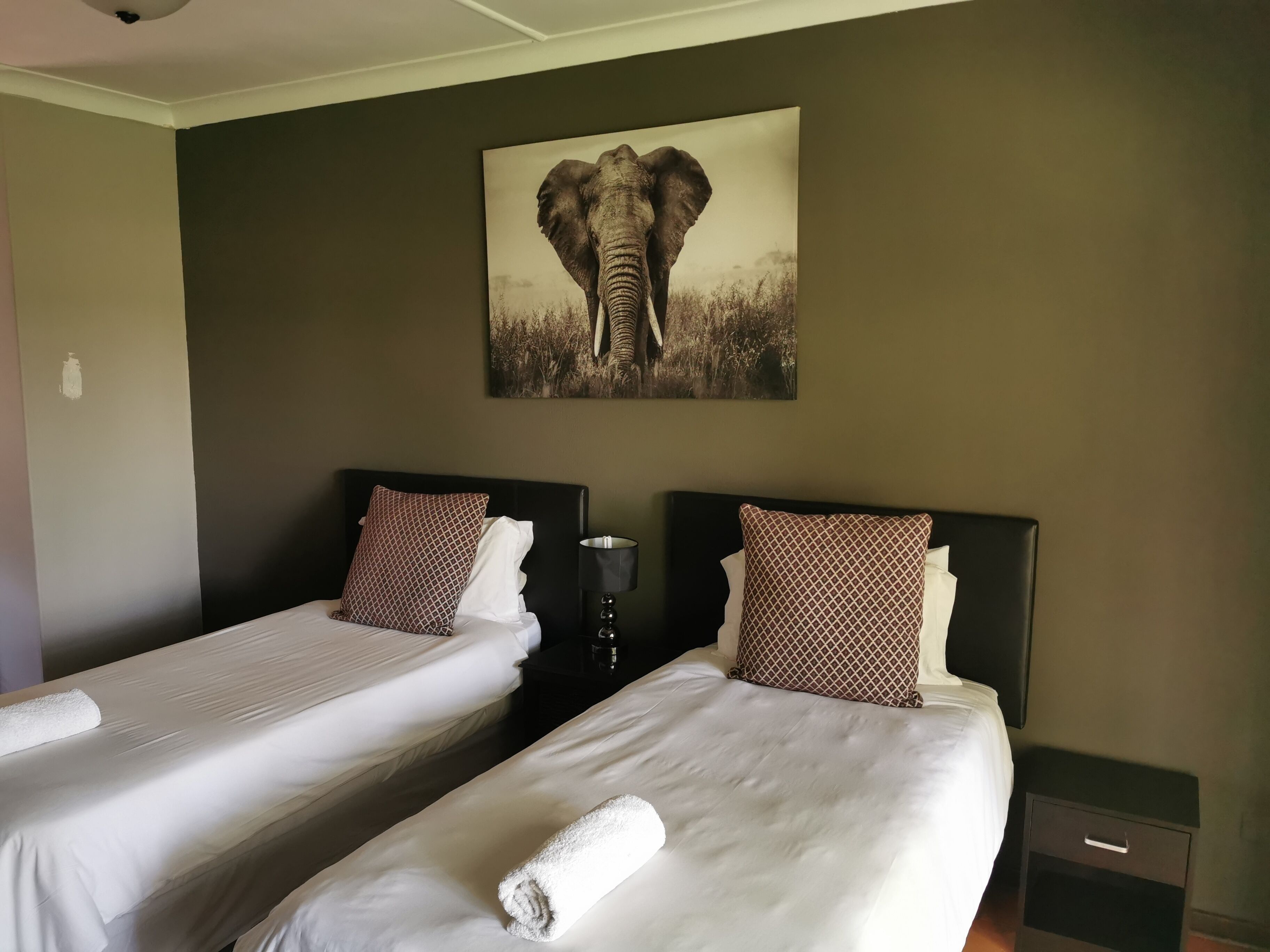 A hotel room with two beds, each with white linens and patterned pillows. There is a painting of an elephant on the wall above the beds.