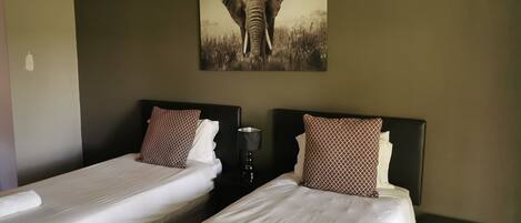 A hotel room with two beds, each with white linens and patterned pillows. There is a painting of an elephant on the wall above the beds.