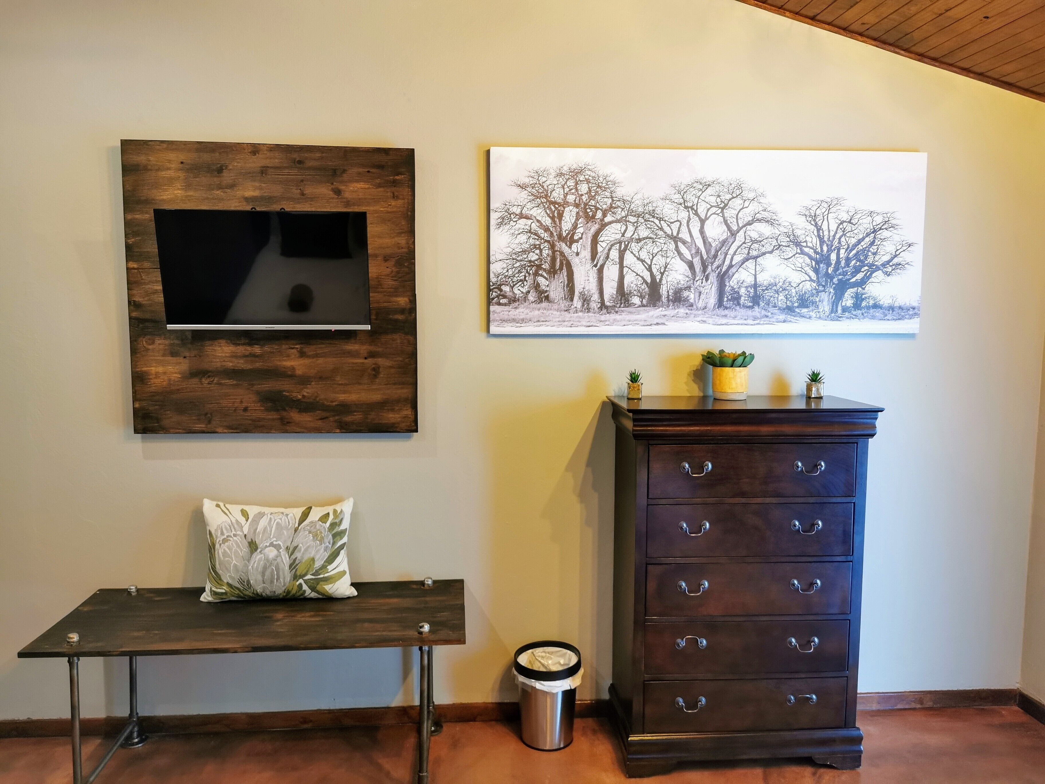 Een kamer met een houten tv-meubel, een bankje met een decoratief kussen, een donkere houten dressoir met meerdere laden, en een prullenbak.