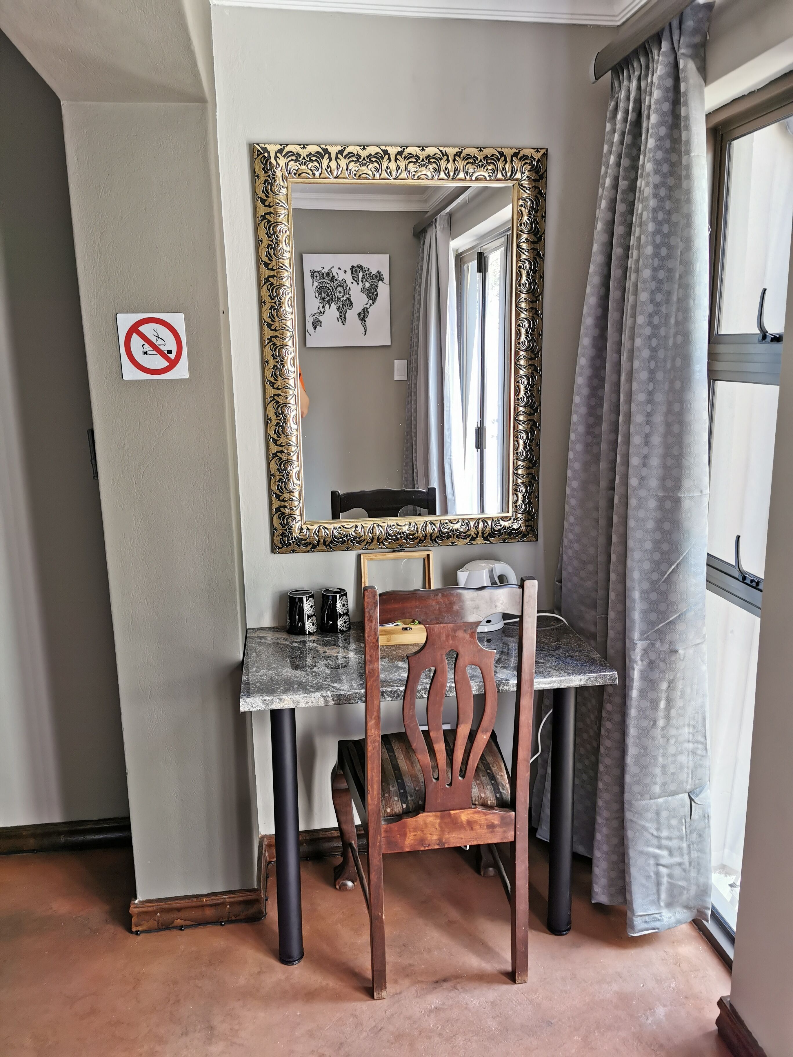 Una habitación pequeña y compacta con escritorio, silla y un espejo grande. Hay un mapa mundial en la pared, un cartel de prohibido fumar y una ventana con cortinas.
