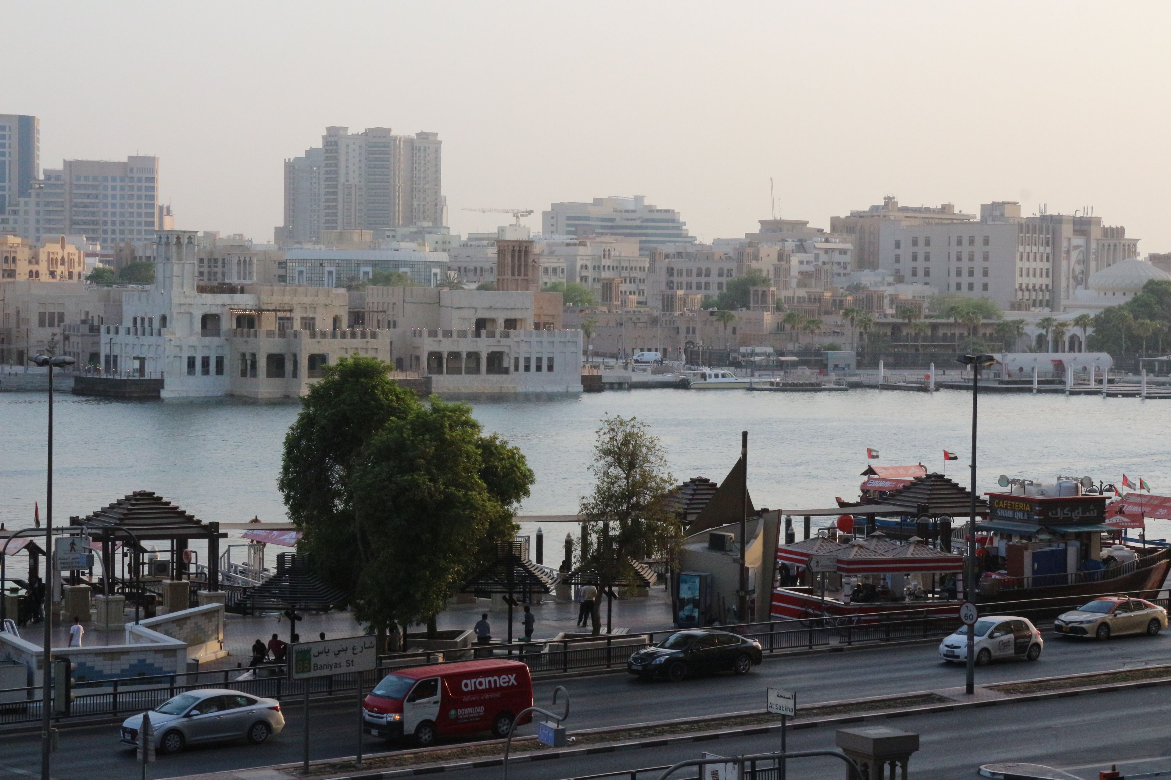 A waterfront cityscape with buildings, a river, and a pedestrian walkway.
